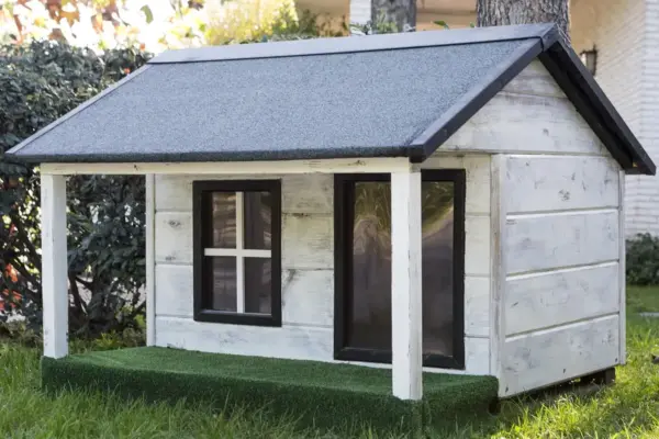Caseta de madera blanca para perro con tejado a dos aguas y porche, sobre el césped de un jardín
